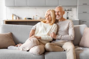 The image depicts a mature couple, potentially discussing their future, including financial planning aspects like a lifetime mortgage. Seated comfortably together on a light gray sofa in their modern, well-lit living room, the setting includes a kitchen in the background. The woman, dressed in a light cream sweater and beige pants, leans towards the man who is similarly dressed in a light gray sweater and pants. Both appear relaxed and content, symbolizing stability and comfort, which might be supported by sound financial planning such as a lifetime mortgage.