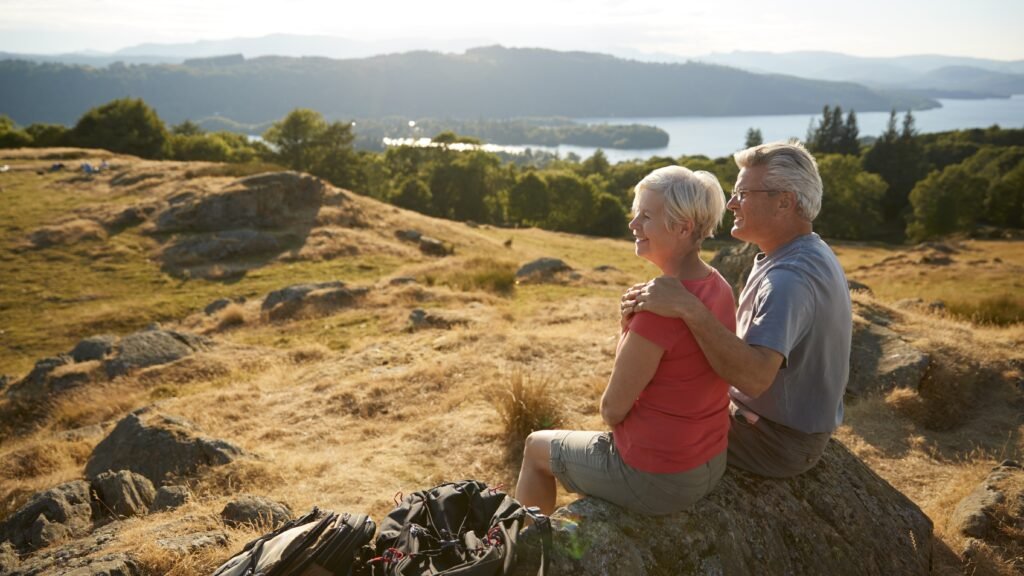 Senior Couple Resting At Top Of Hill On Hike Through Countryside In Lake District UK