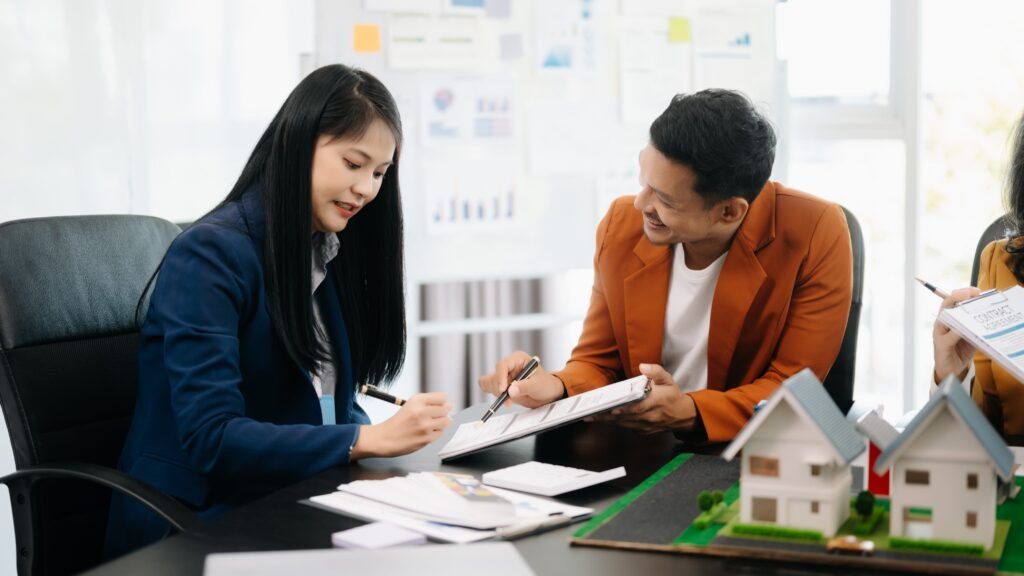 This image shows a professional setting where two people, a woman and a man, are engaged in a business discussion. The woman, dressed in a navy blue blazer, is focused on writing notes on a clipboard. Sitting across from her, the man, wearing an orange blazer, is smiling and pointing towards the documents, suggesting a lively exchange of ideas.